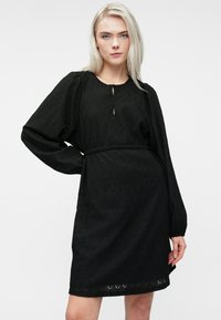 Ξανθιά γυναίκα ντυμένη με μια μαύρη υφασμάτινη μακρυμάνικη φόρεμα με λεπτή ζώνη, στέκεται με το ένα χέρι στη μέση της σε απλό φόντο.