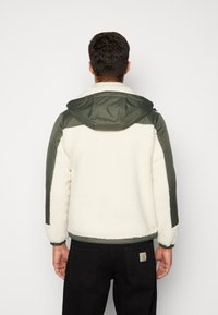 Fleecejacke mit cremefarbenem Körper, grünen Nylon-Akzenten, Kapuze und elastischen Bündchen. Strukturierte Oberfläche mit sicherem Reißverschluss.