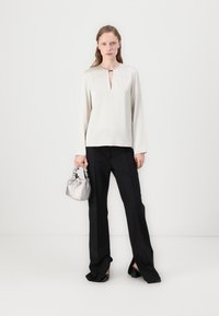 Crème langemouwen top met een sleutelgatdetail aan de voorkant, gecombineerd met zwarte wijde broek en een kleine zilveren satijnen handtas.