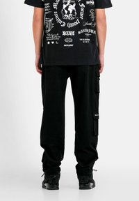 Schwarze Cargo-Hosen mit mehreren Taschen, kombiniert mit einem schwarzen Grafik-T-Shirt, das weiße Designs und Text zeigt. Die Schuhe sind schwarze Sneakers.