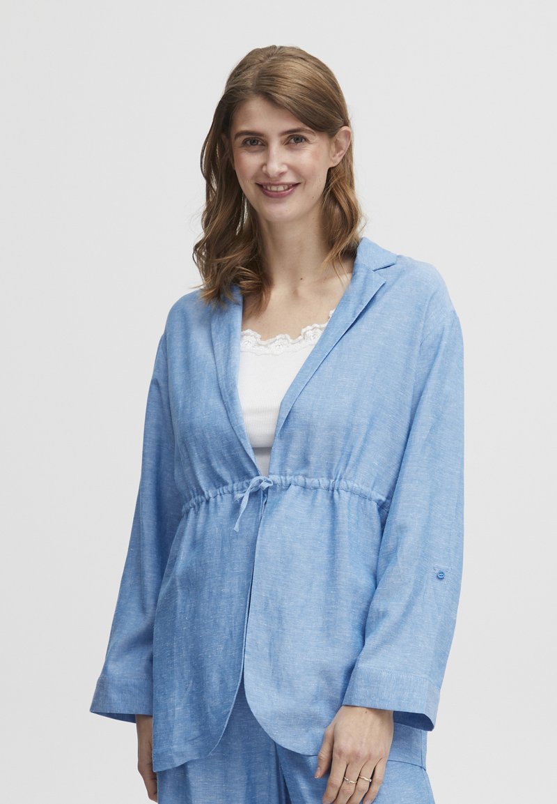 Gaiši zila, garām piedurknēm jaka ar brīvu piegriezumu. Iezīmēta ar elastīgu jostas saiti, apkakles jostu un smalku tekstūru. Nēsāta virs baltas blūzes.