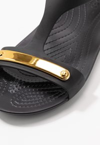 Mustassa kumipohjallisessa sandaalissa on kultasävyinen metallikoriste ylhäällä, teksturoitu sisäpohja, jossa on kohollaan olevia kuvioita, ja sileät reunat.