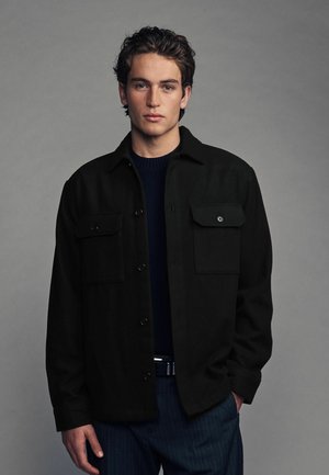 Schwarze Hemdbluse aus einem dicken, strukturierten Material mit zwei Brusttaschen. Über einem dunklen Pullover getragen, kombiniert mit schmal gestreiften Hosen.