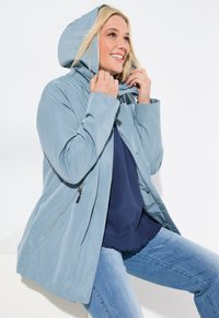 Ljusblå vattentät jacka med huva, med dragkedjestängning och sidofickor. Slät textur, avslappnad design och lös passform.