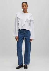 Weißes Langarmoberteil mit Rüschendetails auf einer Seite, kombiniert mit hoch taillierten blauen Jeans und schwarzen flachen Schuhen. Einfaches Design, glatte Textur.