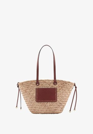 Bolso tote tejido de paja con dos asas largas de cuero marrón, un bolsillo frontal de cuero marrón con tachuelas doradas y cordones laterales ajustables.