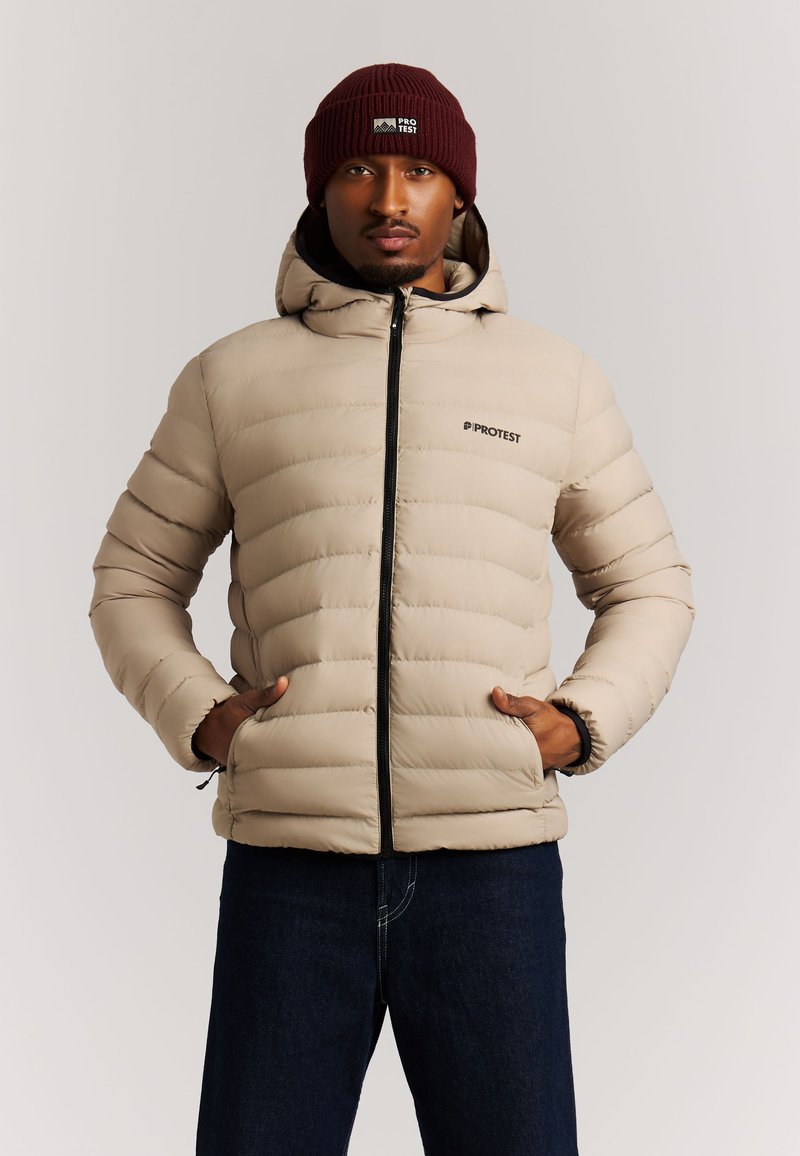 Homme portant une veste matelassée beige avec capuche et un bonnet bordeaux, mains dans les poches, debout devant un fond clair uni.
