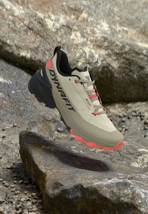Zapatilla de trail running con detalles en beige y coral, suela negra, logo "DYNAFIT", flotando sobre un terreno rocoso.
