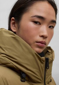 Chaqueta de plumas color caqui con un alto cuello, que presenta un tejido texturizado, un cierre de cremallera y detalles de hardware ajustables en los laterales.