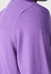 Närbild av en person som bär en lila stickad tröja som visar detaljerad sömnad på axeln och övre ryggen.