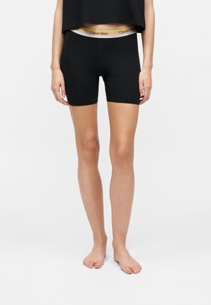 Femme portant un short de cycliste noir avec une ceinture visible de la marque Calvin Klein, debout pieds nus devant un fond uni.