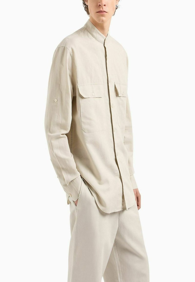 Camicia beige a maniche lunghe con colletto alla mandarino, chiusura frontale a bottoni e due tasche sul petto. Tessuto liscio con vestibilità su misura.
