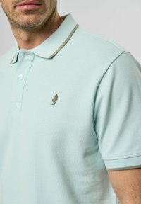 Ljusblå polo med en texturerad väv, som har en kontrasterande brun krage och en liten broderad logotyp på bröstet.