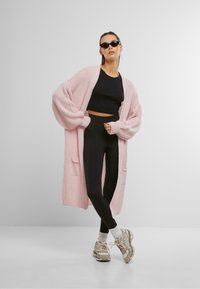 Lichtroze gebreide cardigan met oversized mouwen, zwarte cropped top, zwarte high-waisted legging en beige sneakers met witte sokken.