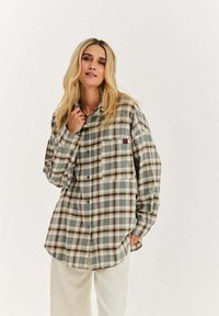 Kobieta z długimi blond włosami, ubrana w luźną koszulę w brązowo-szarą kratę na guziki oraz jasne spodnie, na jednolitym tle.