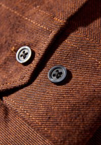 Brązowa tkanina o diagonalnej, prążkowanej fakturze, z dwoma czarnymi guzikami z czterema dziurkami, z widocznym precyzyjnym przeszyciem w okolicy guzików.