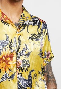 Koszula krótki rękaw w żółtym kolorze z kwiatowym wzorem, zawierająca czarne i różowe motywy roślinne. Gładki materiał o subtelnym połysku i zapięcie na guziki.