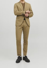 Khakifärgad kostym med skräddarsydd jacka och slim-fit byxor, kombinerad med svarta skor. Slät tyg, minimala detaljer och en modern silhuett.