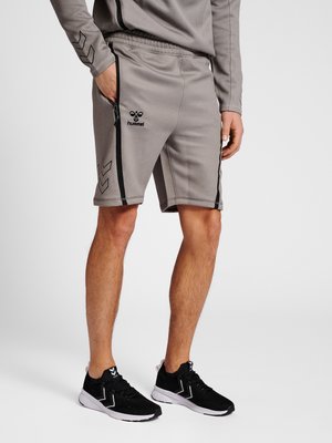 Mand iført grå hummel sportsshorts med sorte striber og sorte sportssko, stående med den ene hånd i lommen.