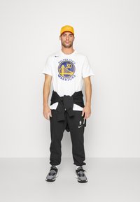 Hvid bomulds t-shirt med "Golden State Warriors" logo, sorte sweatpants med Nike logo, grå sneakers og orange baseball cap.