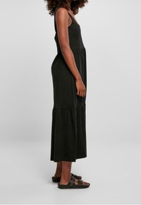 Černé maxi šaty s přiléhavým korzetem a sukní ve více vrstvách. Vyrobené z měkkého materiálu, mají široká ramínka a uvolněný střih. Na nohou má černé sandály.