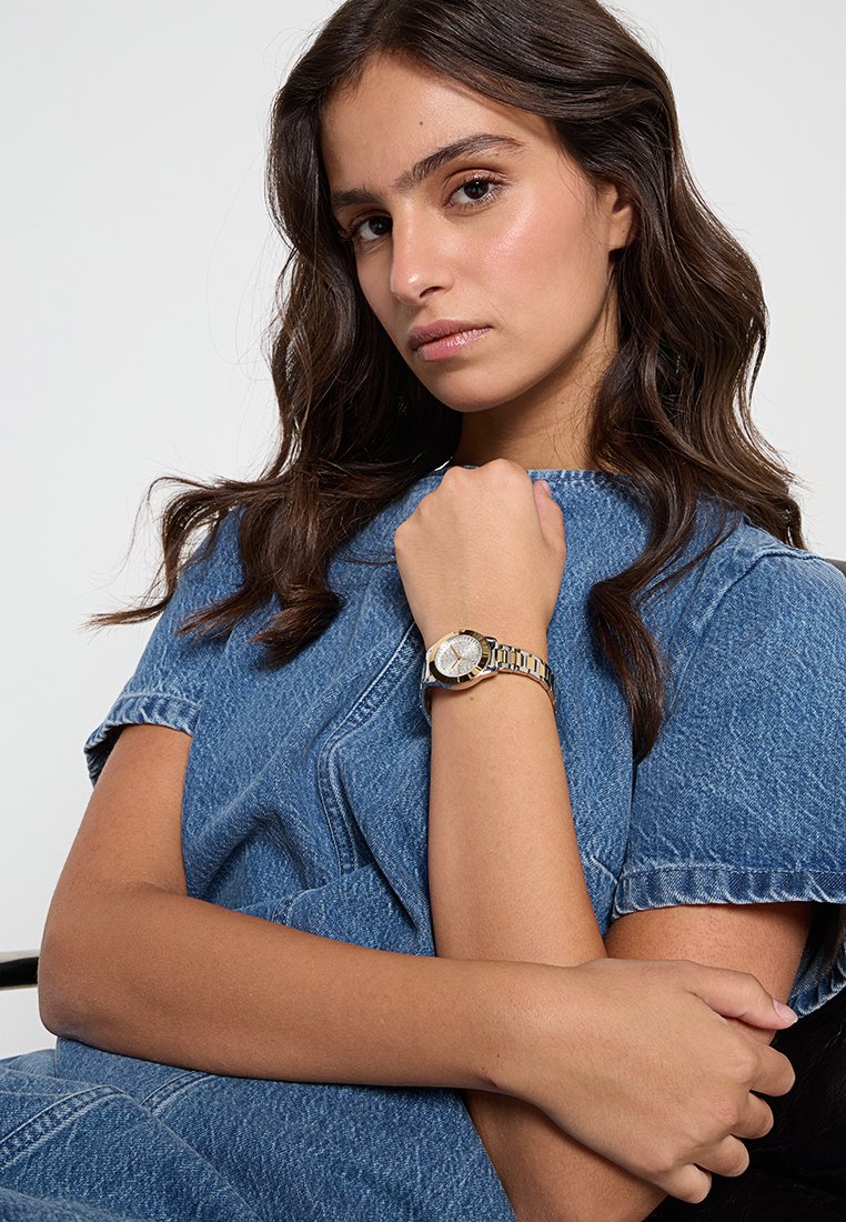 Femme aux longs cheveux noirs portant une tenue en denim bleu, reposant un bras sur l'autre et montrant une montre-bracelet en argent et or.