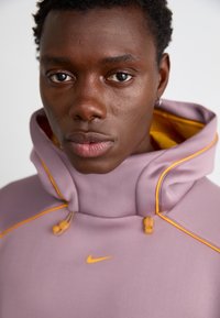 Lila huvtröja med orange detaljer, dragsko och Nike-logotyp på bröstet, med en slät textur och hög krage.