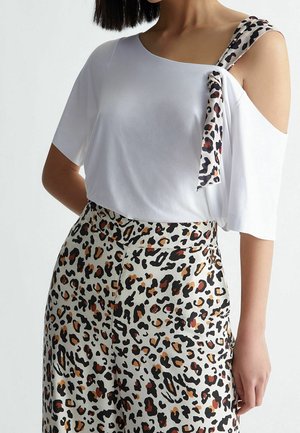 Kobieta w białym topie odsłaniającym ramiona z paskiem w cętki lamparta wiązanym na jednym ramieniu oraz dopasowanych, wysokich spodni w lamparcie wzory.