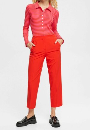 Pantaloni rossi a vita alta con cintura elastica, abbinati a una camicia a maniche lunghe rosa e bianca a righe, con colletto e bottoni. Mocassini neri.