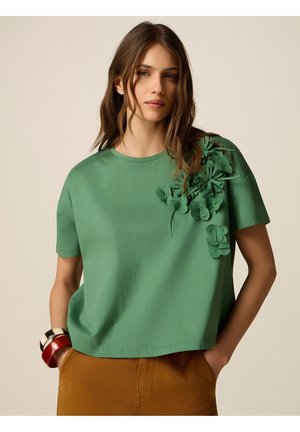Donna con una maglietta verde a maniche corte con fiori di stoffa sulla spalla, pantaloni marroni e braccialetti spessi su uno sfondo beige uniforme.
