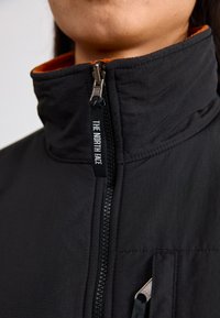 Giacca nera con colletto alto, dotata di una fodera arancione a contrasto e di una cerniera con il marchio "THE NORTH FACE". Texture liscia, design minimal.