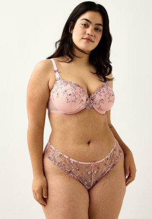 Pitkähiuksinen nainen mallintaa vaaleanpunaista kukkakuosista pitsirintaliivit ja niihin sopivat alushousut, seisten yksinkertaista vaaleaa taustaa vasten.