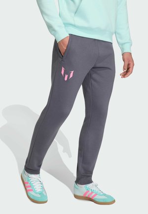 Osoba ubrana w ciemnoszare spodnie dresowe z różowym logo, jasno-niebieską bluzę i turkusowo-różowe buty Adidas stojąca z jedną ręką w kieszeni.