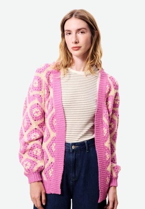 Jeune femme portant un cardigan tricoté à motifs rose et crème sur une chemise rayée blanche et noire, ainsi qu'un jean bleu foncé.