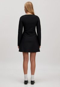 Schwarzes Langarmkleid mit tailliertem Oberteil und ausgestelltem Rock, ausgestattet mit einem geraden Rückenausschnitt und glatter Textur. Tragen mit schwarzen Schuhen und grauen Socken.
