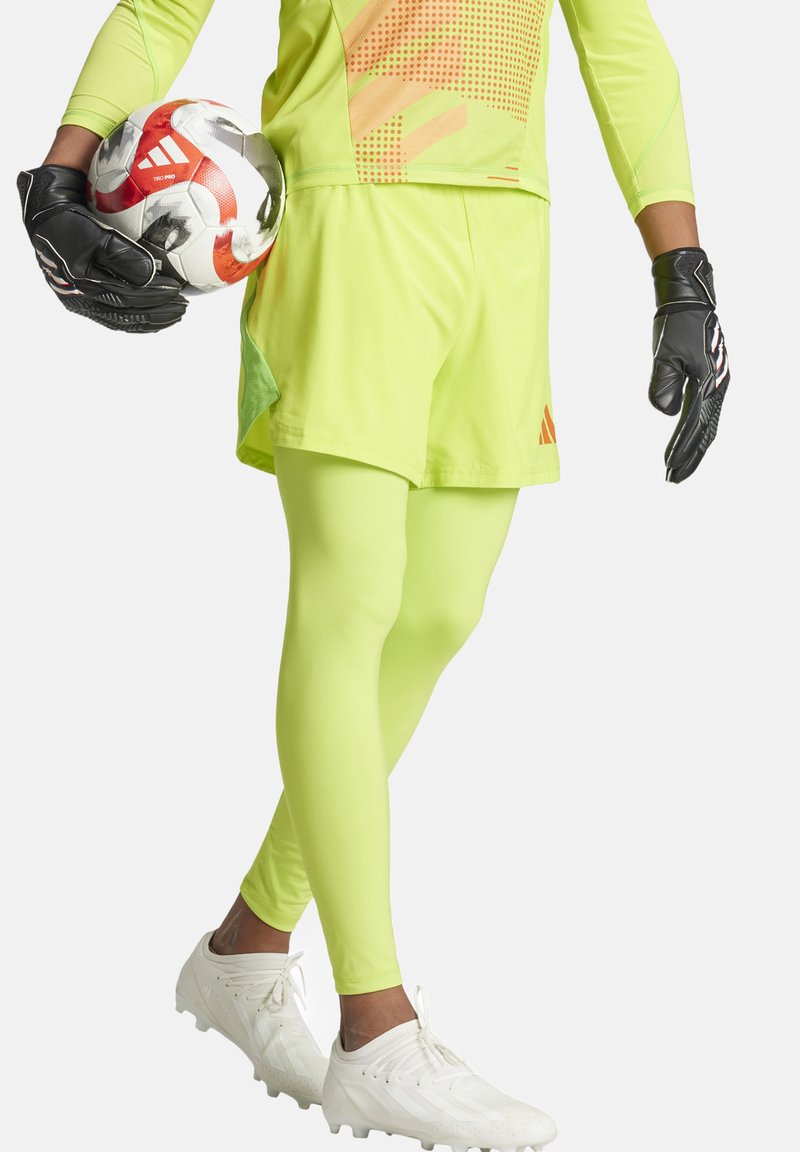 Neon gele sporttrui en -shorts met netaccenten, gecombineerd met bijpassende legging en witte voetbalschoenen. Zwarte handschoenen houden een voetbal vast.