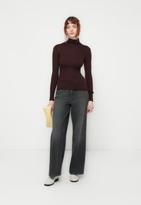 Burgunderfarbener, gerippter Rollkragenpullover kombiniert mit lockeren, grauen weiten Jeans. Die Accessoires umfassen eine pastellgelbe Clutch und cremefarbene Ankle Boots.