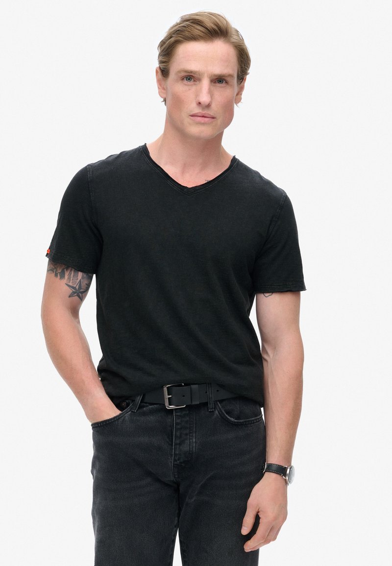 Sort v-hals t-shirt med korte ærmer, lavet af blødt stof. Bæres med mørke jeans og et sort bælte. Har synlige syninger og en tatovering.