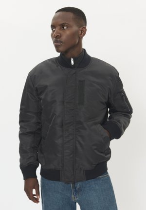 Schwarze Bomberjacke mit einem Reißverschluss, gerippten Bündchen und Taschen. Hergestellt aus glattem Stoff, mit einem leichten Glanz und einer taillierten Passform.