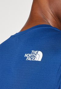 Μπλε κοντομάνικη μπλάζα με υφαντό ύφασμα και λευκό λογότυπο "The North Face" στο πίσω μέρος κοντά στη λαιμόκοψη.