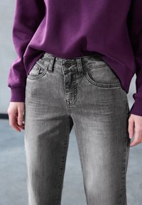 Graue Denim-Jeans mit einer leichten Ausbleichung, Fünf-Taschen-Design und einem schwarzen Knopfverschluss. Kombiniert mit einem lila Sweatshirt.