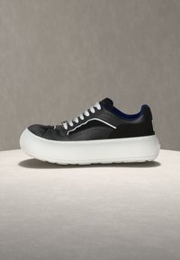 Juodi ir mėlyni sportbačiai su baltais raišteliais ir perdėm stora balta padais neutraliame paviršiuje ir fone.