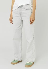 Jeans en denim gris clair à jambes larges, présentant un design classique à cinq poches et une taille haute. Portés avec des baskets vertes.