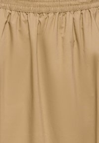 Tissu beige avec une ceinture élastique froncée, une texture lisse et des plis verticaux créant une apparence fluide.