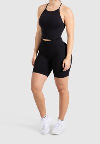 Czarny sportowy top typu crop z cienkimi ramiączkami, połączony z czarnymi rowerowymi szortami o wysokim stanie. Model nosi białe buty sportowe. Gładka faktura materiału.