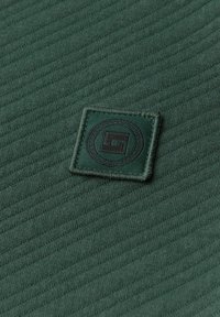 Un tessuto verde scuro presenta un motivo testurizzato con righe diagonali. Al centro si trova una toppa ricamata a forma quadrata con un design circolare.
