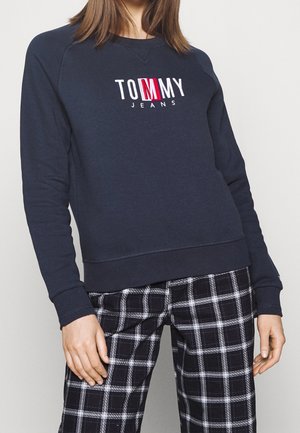 Osoba ubrana w granatową bluzę z logo "Tommy Jeans" oraz czarno-białe spodnie w kratę, stojąca na jednolitym jasnym tle.