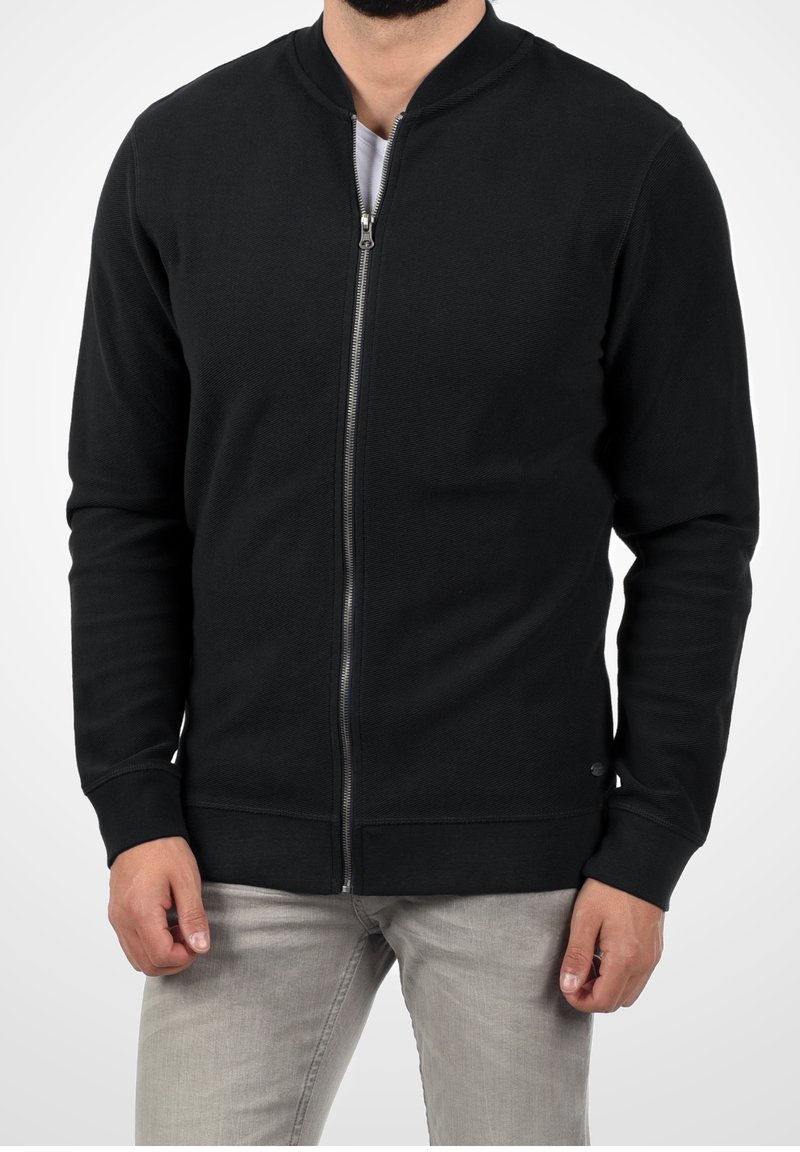 Schwarze Zip-Up-Sweatjacke mit geripptem Stoff, rundem Kragen und Seitentaschen. Verfügt über einen glatten Reißverschluss und enganliegende Bündchen.
