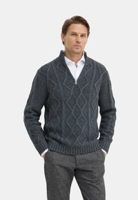 Maglione grigio scuro a maglia con colletto a zip, caratterizzato da un motivo a cavo a rombi e polsini a costine. Indossato sopra una camicia bianca.