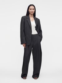 Femme aux longs cheveux foncés portant un costume à rayures sombre oversize et un haut blanc, debout avec une main dans la poche et des sandales sur fond blanc.